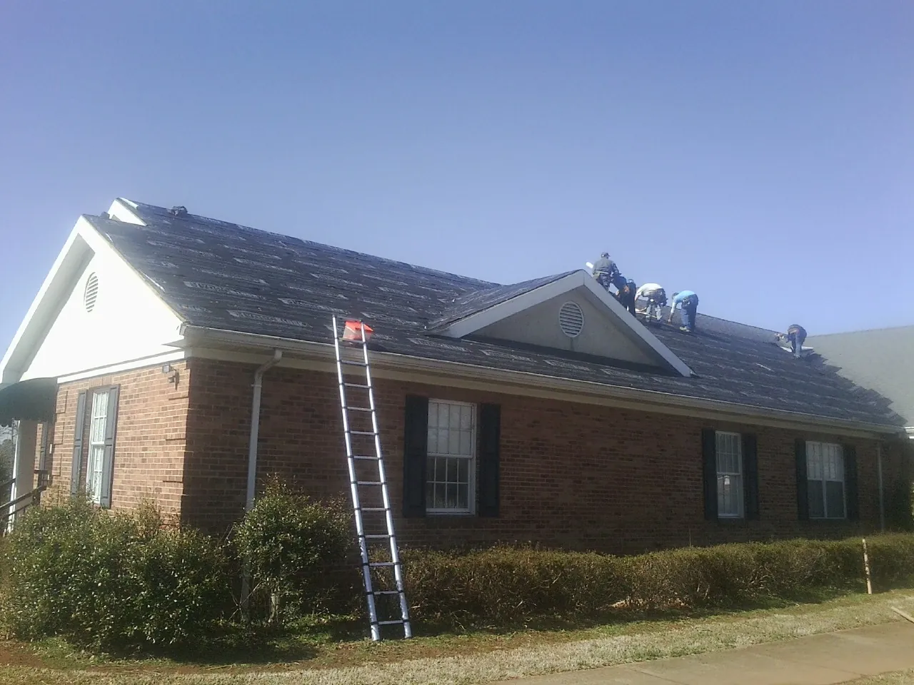 Crew tearing shingles off a brick ranch roof with a ladder set — Emergency Roof Repair work by our crew in Hanska