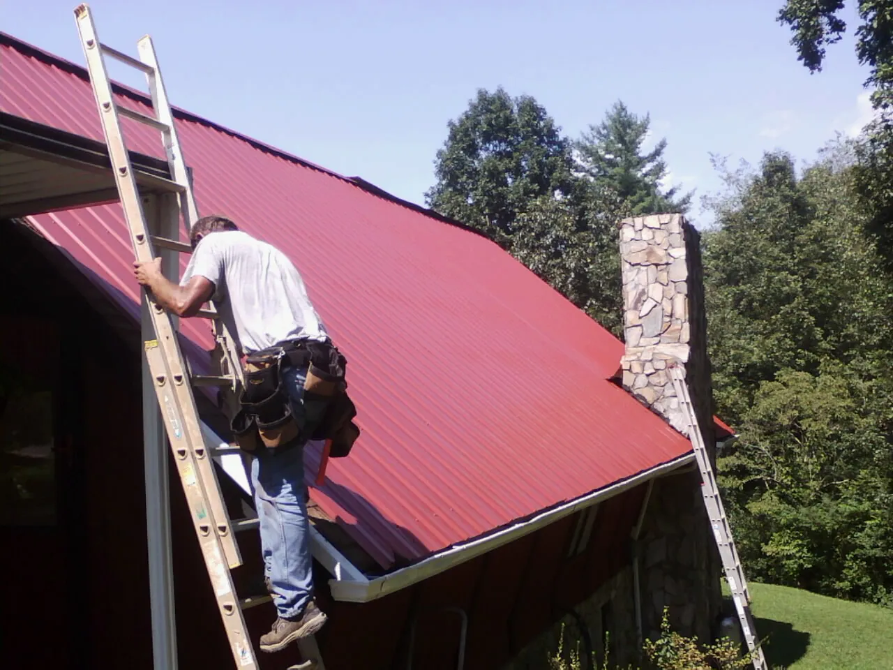 Roofer on a ladder beside a red metal roof and stone chimney — Soffit & Fascia Repair by Roofing Brothers in Hanska
